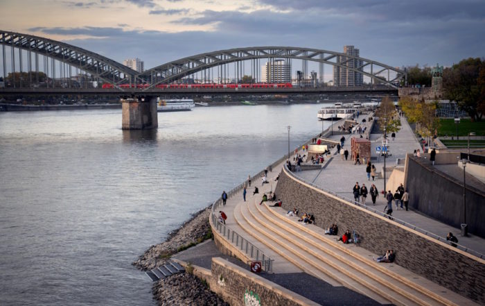 Tour-Reise in Köln: Eine malerische Brücke spannt sich über einen ruhigen Fluss, umgeben von üppigem Grün und klarem blauem Himmel.