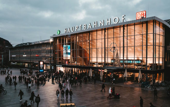 Tour-Reise in Köln: Menschen spazieren um ein großes Gebäude mit einem auffälligen „Bahnhof“-Schild.