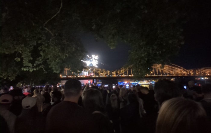 Kölner Lichter: Eine Menschenmenge versammelt sich nachts am Flussufer, mit einer hell beleuchteten Brücke im Hintergrund.