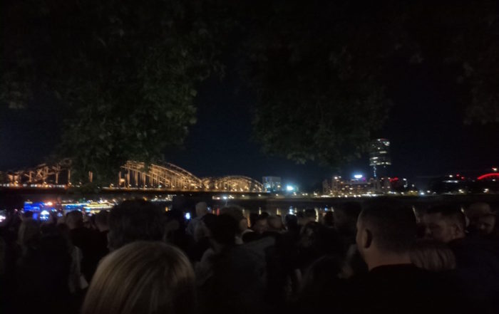 Kölner Lichter: Die Hohenzollernbrücke wird nachts beleuchtet, während eine Menschenmenge vom Flussufer aus zuschaut.