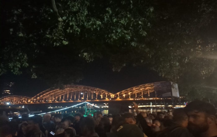 Kölner Lichter: Eine Menschenmenge steht in der Nähe des Wassers, mit einem beleuchteten Schiff und einer Brücke bei Nacht.