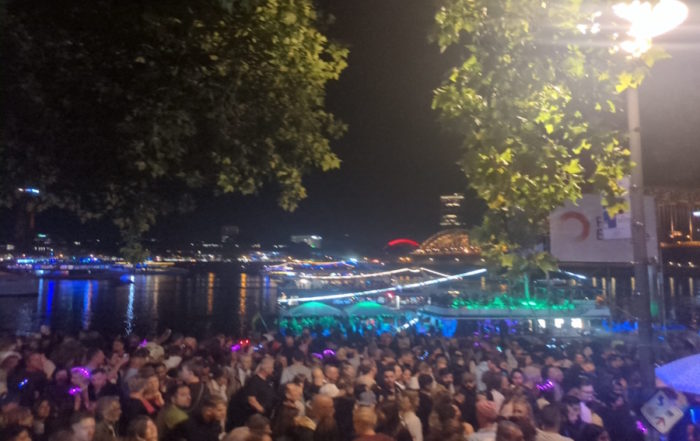 Kölner Lichter: Eine Menschenmenge steht nachts am Fluss und beobachtet ein Schiff und die beleuchtete Hohenzollernbrücke.